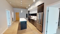 This contemporary urban residence showcases a thoughtfully designed single-floor layout with a distinctive industrial-modern aesthetic. The open-plan kitchen serves as a central highlight, featuring dark wood cabinetry paired with white upper cabinets, stainless steel appliances including a refrigerator, range, and built-in microwave, a clean white countertop, and a freestanding island with a contrasting teal-toned base. Exposed concrete ceilings throughout the living and kitchen areas add an industrial character, complemented by light wood-tone flooring and recessed lighting with pendant fixtures over the island. The bedrooms offer plush carpeting, generous sliding-door closets, and large black-framed windows that frame expansive urban skyline views, filling each room with abundant natural light. Two bathrooms provide polished functionality: one features a glass-enclosed walk-in shower with a wide vanity and large mirror, while the second includes a soaking tub with overhead shower, tile surround, and a matching dark wood vanity with a white countertop. A slatted wall accent panel in the living area adds a modern design touch. Throughout, the neutral color palette, clean lines, and quality finishes create a cohesive, contemporary ambiance well-suited to urban living.