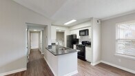 This single-story residence offers a well-organized layout with a variety of thoughtfully designed spaces throughout. The open-concept kitchen serves as a functional centerpiece, featuring white cabinetry, dark countertops, a kitchen island with an integrated sink, and black appliances including a refrigerator, range, and microwave. A dedicated laundry area with a washer is conveniently accessible directly from the kitchen. Hardwood-style flooring runs throughout the main living and kitchen areas, contributing to a clean, cohesive aesthetic.

The living room is a standout space, characterized by soaring vaulted ceilings, a ceiling fan, and a curved bay window arrangement that invites abundant natural light. The open feel is further enhanced by the room's generous proportions and neutral wall tones.

Bedrooms are carpeted for comfort, with at least one featuring vaulted ceilings and a prominent arched window that frames outdoor views. Additional bedrooms are well-proportioned with standard ceilings and natural light from individual windows.

Two bathrooms are present: one with a single-sink vanity, dark countertop, white cabinetry, and a glass-enclosed shower, and a second full bathroom with a tub-shower combination and matching vanity. Both bathrooms feature hardwood-style flooring and neutral finishes throughout.
