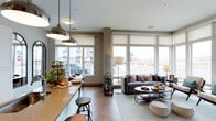 This single-floor space showcases an open-concept layout that thoughtfully connects the kitchen and living areas into one cohesive environment. The kitchen features a long, warm-toned countertop that doubles as a breakfast bar, complete with industrial-style bar stools and pendant lighting overhead. Sleek metallic dome pendant lights hang above the counter, adding a modern, polished touch to the space.

Large floor-to-ceiling windows line the exterior wall, flooding the interior with abundant natural light while offering open views of the surrounding streetscape. Roller shades provide flexible light control without compromising the airy feel. The living area is anchored by a comfortable gray sofa with decorative pillows, positioned around a circular tiered coffee table that sits atop a light-toned area rug. A small bar cart and coordinating floor lamps contribute to the well-appointed, functional arrangement.

Throughout the space, warm wood-toned flooring and neutral gray walls create a cohesive, contemporary palette. Thoughtful styling details, including decorative greenery, books, and curated accessories, enhance the overall inviting and well-organized ambiance of the space.