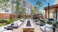 This outdoor amenity space is designed to deliver a resort-like experience within a modern multi-story residential community. The architecture blends grey paneling, warm brick accents, and natural stone detailing to create a cohesive and visually appealing environment throughout.

At the heart of the community is a spacious, crystal-clear swimming pool surrounded by a generous sun deck outfitted with lounge chairs and red market umbrellas, ideal for relaxing in the open air. Adjacent to the pool, a beautifully landscaped courtyard serves as a central gathering hub, featuring a striking elongated fire feature set on a stone base and flanked by contemporary outdoor sofas and lounge chairs with crisp white cushions. Mature trees and curated plantings add greenery and a sense of natural tranquility to the paved gathering areas.

A covered porch area with a warm wood-paneled ceiling provides a shaded retreat furnished with all-weather seating, offering direct views into the courtyard through floor-to-ceiling glass doors and windows. Nearby, a putting green framed by expansive steel-framed glass walls and natural stone columns adds a recreational element to the space. String lights overhead and a pergola structure further enhance the ambiance, making the community equally inviting during evening hours.