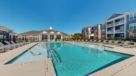Located at 100 Conference Centre Dr in Cartersville, Georgia, this outdoor amenity area is centered around a large, resort-style swimming pool with crystal-clear turquoise water. The pool is generously sized with a shallow entry zone featuring built-in lounge chairs that allow residents to relax partially submerged, as well as traditional stainless steel entry rails along the perimeter. Surrounding the pool is a spacious concrete sundeck lined with rows of chaise lounges, providing ample space for sunbathing and outdoor relaxation. Shaded umbrella seating areas are available for those seeking relief from the sun. The centerpiece of the complex is a well-appointed clubhouse building, designed in a classic Southern architectural style with gabled rooflines, prominent white columns, a chimney, and large windows that allow natural light to fill the interior. A pergola structure extends from one side of the clubhouse, adding a charming outdoor gathering space. The entire pool area is enclosed by a black wrought-iron fence, ensuring both safety and aesthetic appeal. Multi-story residential buildings with brick and siding exteriors frame the backdrop, completing the cohesive community setting. The space benefits from abundant natural light and open sky views, creating a bright and inviting outdoor retreat.