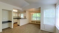 This single-floor space offers an open, connected layout that integrates the kitchen and dining area into one cohesive living environment. The kitchen is well-appointed with white cabinetry featuring light wood accents, dark countertops, and a peninsula that provides additional workspace and serves as a natural divider between the kitchen and the adjacent dining area. A recessed ceiling detail above the kitchen adds subtle architectural definition to the space.

The dining area is highlighted by an elegant chandelier that serves as a focal point, adding a touch of classic charm. A large picture window floods the area with natural light and frames views of the surrounding greenery, creating a bright and inviting atmosphere. Built-in shelving beneath the window offers both functional storage and a cozy seating nook, adding character and practicality to the space.

Neutral tones throughout — including beige carpet, white walls, and light-colored cabinetry — create a clean, versatile backdrop. Additional windows along the adjacent wall further enhance the natural light. The overall ambiance is warm, airy, and well-suited for both everyday living and entertaining.