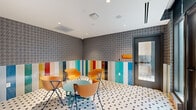This pet spa is a thoughtfully designed, amenity-rich facility that combines bold aesthetics with practical functionality. The main lounge area sets a lively tone with its striking houndstooth-patterned wallpaper on the upper walls and a vivid multicolored vertical stripe wainscoting below. Underfoot, a classic white hexagonal tile floor with charcoal dot accents ties the space together. A cluster of vibrant orange molded chairs surrounding a teal accent table provides a comfortable waiting area for pet owners. Recessed lighting illuminates the space evenly from the white ceiling above. An adjacent grooming and wash room is outfitted with white subway tile walls accented by teal and blue tilework, a deep utility sink with a gooseneck faucet, built-in shelving, and a raised pet wash station — all finished with the same hexagonal tile flooring for a cohesive look. The space also connects to an outdoor turf area enclosed by modern concrete and wood-panel walls, featuring lush artificial grass, raised garden planters, wall-mounted lighting, and a waste station. A blue shade sail overhead and sweeping urban skyline views complete this well-appointed outdoor retreat for pets and their owners.