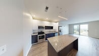 Located in the heart of downtown Fort Wayne, Indiana, this thoughtfully designed 2-bedroom, 2-bathroom residence offers a clean, contemporary aesthetic throughout. The open-concept kitchen and living area serves as the centerpiece of the home, showcasing a two-tone kitchen with crisp white upper cabinets and deep navy lower cabinets, granite countertops, a large island with additional seating space, and a full suite of stainless steel appliances. Wood-look flooring flows throughout the main living areas, complementing the neutral color palette and modern finishes. Recessed lighting and a sleek linear pendant fixture illuminate the kitchen, while large windows and a sliding glass door flood the living area with natural light and provide access to a private balcony with striking elevated views of the downtown Fort Wayne skyline. The spacious primary bedroom features a generously sized closet and large windows offering impressive views of the city's architectural landmarks, including historic church steeples. Both bathrooms are finished with granite vanity tops, dark wood cabinetry, and tile flooring, with one featuring a tub-shower combination. The second bedroom also benefits from abundant natural light. Clean lines, quality materials, and an elevated urban setting define this well-appointed residence.