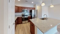 This single-floor residence offers a thoughtfully designed open-concept layout that seamlessly connects the kitchen and living areas. The kitchen is a standout feature, showcasing rich cherry wood cabinetry, light granite countertops, a large center island with a built-in sink, stainless steel appliances including a gas range, built-in microwave, and French door refrigerator, all set atop light hardwood-style flooring. Pendant lighting accents the island, adding warmth to the space. The open living area extends from the kitchen, featuring the same light hardwood-style flooring, a ceiling fan, and sliding glass doors that invite natural light and provide access to an outdoor area. The bedrooms are carpeted and generously sized, each benefiting from multiple windows that allow abundant natural light. At least one bedroom includes a dedicated walk-in closet with built-in shelving. The bathrooms are finished with granite-topped white vanities, framed mirrors, and hardwood-style flooring. One bathroom features a glass-enclosed walk-in shower, while another is equipped with a full vanity and toilet. The unit's neutral warm-beige palette throughout creates a clean, cohesive, and move-in-ready ambiance.