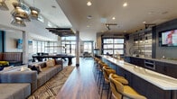 Located at 22004 E Quincy Ave in Aurora, Colorado, this single-floor community amenity space is thoughtfully designed to encourage socializing and relaxation. The interior combines modern industrial aesthetics with warm, inviting finishes, resulting in a cohesive and upscale atmosphere throughout.

The open-concept layout features rich hardwood-style flooring that flows seamlessly across the space. A striking lounge area anchors one section of the room, highlighted by a floor-to-ceiling reclaimed wood fireplace surround set against deep teal walls, flanked by tufted banquette seating and stylish accent tables with gold finishes. Geometric patterned rugs add visual texture beneath the carefully curated furniture arrangements.

A long bar counter with a white quartz surface and navy cabinetry runs along one side, lined with curved amber-upholstered barstools. The adjacent fully equipped kitchen features stainless steel appliances, dual-basin sinks, pendant lighting, and a wall-mounted television, making it ideal for catered events or communal cooking.

The game room area boasts soaring ceilings with dramatic angular beam details, a full-size billiards table, foosball, and a wall-mounted TV. Expansive floor-to-ceiling windows and glass garage-style doors flood the space with natural light and provide direct views of the outdoor patio, seamlessly connecting the interior and exterior environments.