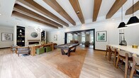Located at 100 Leslie Oaks Dr in Lithonia, Georgia, this single-story community clubhouse offers a generously sized, thoughtfully designed gathering space that blends modern aesthetics with warm, inviting details. The entrance greets visitors with a striking black-and-white checkered tile floor beneath soaring ceilings, anchored by a large, framed landscape painting that commands attention upon arrival. A reception counter and branded welcome mat set a polished, professional tone from the moment you step inside. The main living area transitions seamlessly from the foyer, featuring wide-plank wood-look flooring, exposed dark wood ceiling beams, and an abundance of natural light streaming through multiple large windows that frame lush greenery outside. A comfortable lounge zone is anchored by a caramel leather sectional sofa, a stone-surround fireplace, and built-in shelving with curated décor. A full-size pool table serves as a central entertainment focal point, while a bar-height dining table with stylish seating offers a social gathering spot. Toward the rear, a kitchenette with wood cabinetry, pendant lighting, and modern appliances supports events and everyday use. Throughout, the space is accented by statement light fixtures, curated artwork, and lush indoor plants, creating a refined yet relaxed ambiance suited for community connection and recreation.