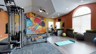 This well-appointed fitness center delivers a dynamic workout environment designed to motivate and inspire. The space is immediately distinguished by a bold, large-scale abstract mural rendered in vivid reds, blues, and yellows, which commands attention and sets an energetic tone throughout the room. Warm terracotta-orange accent walls complement the artwork and create a visually stimulating atmosphere.

The flooring transitions between durable hardwood-style planking and patterned carpet tiles, thoughtfully zoning the space into distinct workout areas. The cardio section features elliptical trainers and step machines positioned near bright windows that allow natural light to fill the room. A cable and strength machine station occupies one side, offering a range of resistance-based exercises, while a rowing bench adds to the functional training options.

A dedicated stretching and yoga area with a teal exercise mat provides a calm counterpart to the more intensive equipment zones. Decorative planters with lush greenery add a refreshing natural touch, while a mounted flat-screen television and ceiling fan ensure comfort during extended sessions. Recessed lighting and an arched window further enhance the bright, welcoming ambiance of this thoughtfully curated fitness space.