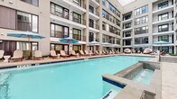 This contemporary multi-story residential complex is designed around an impressive outdoor amenity corridor that seamlessly blends indoor and outdoor living. At its centerpiece is a resort-style swimming pool framed by a generous pool deck lined with lounge chairs and blue-umbrella shaded seating, creating a relaxed, resort-like atmosphere within the building's inner courtyard. The surrounding multi-level structure features private balconies, large framed windows, and a neutral façade with modern architectural detailing.

An expansive covered terrace extends along one side of the complex, finished with large-format stone-look tile flooring and supported by bold structural columns. Floor-to-ceiling glass walls allow natural light to flood interior spaces while offering panoramic views of the surrounding cityscape. The terrace incorporates an outdoor kitchen and bar area with striking stacked-stone cladding, complemented by string lighting and multiple seating configurations for both casual lounging and social gatherings.

Adjacent to the pool, a spa area features contemporary water loungers and pod-style seating, adding a boutique hotel aesthetic to the amenity space. The open-air design encourages natural ventilation while the covered pergola structures provide shade and shelter. Together, these amenity spaces deliver a cohesive, upscale outdoor experience tailored for community living.