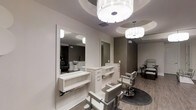 This single-floor salon space is thoughtfully designed to deliver a refined, upscale experience for clients and professionals alike. The interior presents a cohesive neutral palette of soft whites, warm grays, and taupe tones, creating a calm and sophisticated ambiance throughout. Multiple styling stations line the walls, each outfitted with sleek white vanity units, generous mirror panels framed in clean white molding, and dedicated drawer storage to keep tools organized and accessible. Classic adjustable styling chairs in complementary gray upholstery are positioned at each station, balancing functionality with aesthetic appeal. The flooring features light wood-tone planks that add warmth and visual continuity across the space. Overhead, decorative drum pendant lights with a contemporary crystal-inspired design serve as standout fixtures, casting a bright, even glow across the work areas. Ceiling details include subtle recessed tray accents that add architectural dimension. Toward the rear, a textured accent wall in a tonal geometric pattern introduces visual depth and a touch of luxury. Additional pedicure stations are visible in the background, suggesting a full-service salon environment. The overall design is clean, modern, and carefully curated for a professional setting.