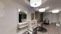 This single-floor salon space is thoughtfully designed to deliver a refined, upscale experience for clients and professionals alike. The interior presents a cohesive neutral palette of soft whites, warm grays, and taupe tones, creating a calm and sophisticated ambiance throughout. Multiple styling stations line the walls, each outfitted with sleek white vanity units, generous mirror panels framed in clean white molding, and dedicated drawer storage to keep tools organized and accessible. Classic adjustable styling chairs in complementary gray upholstery are positioned at each station, balancing functionality with aesthetic appeal. The flooring features light wood-tone planks that add warmth and visual continuity across the space. Overhead, decorative drum pendant lights with a contemporary crystal-inspired design serve as standout fixtures, casting a bright, even glow across the work areas. Ceiling details include subtle recessed tray accents that add architectural dimension. Toward the rear, a textured accent wall in a tonal geometric pattern introduces visual depth and a touch of luxury. Additional pedicure stations are visible in the background, suggesting a full-service salon environment. The overall design is clean, modern, and carefully curated for a professional setting.