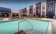 The outdoor amenity level at Ely Apartments is a thoughtfully designed resort-style courtyard that serves as the social hub of the community. At its center is a large, sparkling swimming pool with crystal-clear water, complemented by a separate spa tucked along the building's edge. The pool deck is generously sized, offering ample space for lounging on blue and pink chaise chairs arranged around the water. Private cabanas with slatted wood privacy screens, striped canopies, and tropical-print cushions provide shaded retreats for residents seeking a more secluded outdoor experience. A pergola-covered lounge area features an outdoor kitchen with built-in grilling stations, a mounted television, and comfortable seating, making it an ideal gathering spot for entertaining. Concrete walkways wind through the courtyard, bordered by low-water desert landscaping featuring ornamental grasses, shrubs, and gravel ground cover well-suited to the arid climate. The complex is enclosed by sleek black iron fencing with masonry pillar entries, ensuring privacy and security. The multi-story residential building surrounds the courtyard on multiple sides, with resident balconies overlooking the amenity space. The modern exterior features a bold blue, gray, and white color palette with large windows throughout.