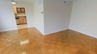 This single-floor space offers a clean, functional layout designed for comfortable everyday living. The main living area is finished with classic parquet hardwood flooring in a warm honey tone, lending a timeless character to the interior. Neutral, light-colored walls throughout create a bright and airy atmosphere, providing a versatile backdrop for a variety of furnishing styles. A low partial wall or architectural divider defines the space, adding subtle structure without sacrificing the open feel of the floor plan. Ceiling-mounted HVAC vents ensure year-round climate comfort throughout the living area. Transitioning from the main room, the adjacent kitchen is visible through an open passageway and features wood-toned cabinetry, light countertops, and tile flooring, offering a practical and well-equipped cooking space. The seamless flow between the living area and kitchen makes the layout especially well-suited for those who value connected, open-concept living. Overall, the space presents a tidy, move-in-ready interior with consistent finishes and a welcoming, straightforward design.