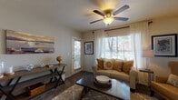 This single-floor residence offers a thoughtfully designed layout that balances comfort and functionality across its key living areas. The living room serves as the heart of the home, featuring plush carpeting, a ceiling fan with integrated lighting, and generous windows dressed with sheer white curtains that invite abundant natural light. Views of a landscaped outdoor area and pool are visible through the windows and a private exterior door, enhancing the sense of connection to the outdoors. Seating is ample, with a cushioned sofa and warm golden accent chairs arranged around a patterned area rug and dark wood coffee table. The dining area, situated adjacent to the kitchen, accommodates a round table with four chairs beneath angled ceilings and tasteful wall art. The kitchen is efficiently arranged with white cabinetry, full-size appliances including a refrigerator, electric range, built-in microwave, and dishwasher, all set over tile flooring. The bedrooms are carpeted and well-lit by natural light through curtained windows, with one bedroom featuring a bold accent wall and a walk-in closet. The bathroom offers a vanity with ample counter space, tile flooring, and a shower-tub combination, completing this comfortable and cohesive living space.