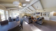 This single-floor space is defined by its dramatic vaulted ceiling adorned with white-painted exposed beams and multiple skylights that flood the interior with abundant natural light. Ceiling fans are positioned throughout to ensure comfortable airflow across the expansive open layout. The space is organized into several distinct functional zones that flow seamlessly into one another. A dining area near the entrance features a large round table with upholstered chairs, ideal for group gatherings. The central lounge area is furnished with generous gray sofas arranged around a wooden coffee table, creating a relaxed and inviting atmosphere. A built-in shelving unit and fireplace anchor one wall, flanked by a mounted television — offering both a decorative focal point and a comfortable media viewing area. Additional built-in cabinetry provides ample storage and display space. Toward the rear, a game room with a pool table is visible, separated subtly from the main living space, adding a recreational dimension to the layout. Large windows along the perimeter allow views of the surrounding greenery, bringing the outdoors in. The neutral carpet and warm, layered lighting create a cohesive and welcoming ambiance throughout the entire space.