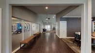 This single-story amenity center is thoughtfully designed to offer residents a comprehensive range of facilities within a cohesive, modern interior. The main corridor welcomes visitors with rich dark hardwood-style flooring, vaulted ceilings with ceiling fans, and a display area lined with glass panels, leading seamlessly into various functional zones.

The fitness center is a standout feature, boasting soaring ceilings with acoustic tile panels and linear lighting, an energizing motivational mural wrapping the upper walls, and an array of cardio equipment including treadmills, ellipticals, and stationary bikes. Clerestory windows bring in abundant natural light while maintaining privacy.

A dedicated pet wash room provides a self-service dog wash station finished with cheerful illustrated wallpaper and easy-clean tile surfaces. Adjacent to this, an indoor hot tub room is lined with large windows overlooking the outdoor pool area, offering a bright and relaxing retreat.

The leasing and business area features a distinctive vaulted wood-plank ceiling with track lighting, herringbone-patterned carpet, a central island countertop, and direct views of the pool through wide glass doors. A comfortable lounge space with high ceilings, plush seating, and framed artwork provides an inviting area for residents to gather and unwind.