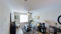 This single-story space centers around a comfortable and thoughtfully designed living room that balances functionality with a welcoming aesthetic. The room features light gray walls that create a neutral, airy backdrop, complemented by smooth white ceilings equipped with a multi-blade ceiling fan with an integrated light fixture, ensuring year-round comfort and ambient lighting. Light wood-toned flooring runs throughout, adding warmth to the space.

A large window fitted with white horizontal blinds allows natural light to fill the room while maintaining privacy. The living area is anchored by a gray sofa adorned with decorative teal, yellow, and patterned throw pillows, paired with a bold black-and-white patterned area rug that adds visual contrast. A rectangular dark-finished coffee table serves as a central focal point, flanked by coordinating end tables with table lamps that provide soft, supplemental lighting.

Wall art in soft teal and yellow tones echoes the color palette of the furnishings, contributing to a cohesive and curated feel. A small accent chair and additional decorative greenery complete the space, adding character and a lived-in warmth to this inviting single-floor layout.