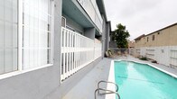 This outdoor space centers around a generously sized rectangular swimming pool with clear, turquoise water, accessible via stainless steel handrails. The pool deck is finished in smooth concrete, providing ample space for movement and relaxation around the perimeter. A collection of lounge chairs and patio seating is arranged near the far end of the pool, offering a dedicated area to unwind and enjoy the surroundings.

The space is flanked on one side by a multi-story building with a modern gray stucco exterior, featuring white lattice-style railings along its walkways and balconies. Large windows line the ground floor, allowing natural light to connect the interior and exterior spaces. On the opposite side, a beige stucco building with decorative signage forms a visual boundary, adding character to the courtyard setting.

A white metal security fence encloses the far end of the pool area, providing safety and a sense of enclosure, while a mature tree adds a touch of greenery to the otherwise urban landscape. The overall ambiance is functional and inviting, designed with communal outdoor living in mind. The layout and amenities make this pool area a practical and appealing gathering space for residents or guests seeking outdoor recreation.
