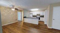 This single-floor space is designed with an open-concept layout that seamlessly connects the kitchen and living areas, creating a functional and cohesive environment. Rich, dark hardwood flooring extends throughout, adding warmth and a sense of continuity across the space.

The kitchen is a standout feature, outfitted with crisp white cabinetry that provides generous storage and a clean, classic aesthetic. Dark countertops offer a striking contrast against the white cabinets, and a central island with a built-in sink serves as both a practical workspace and a natural gathering point. A full suite of appliances, including a refrigerator and range, are integrated into the kitchen layout.

A textured ceiling adds subtle architectural detail overhead, while a ceiling fan with a light fixture ensures comfortable air circulation throughout the open living area. Neutral-toned walls complement the overall color palette, keeping the space feeling bright and inviting.

A hallway visible from the main living area leads to additional rooms, offering a clear and practical flow throughout the floor plan. The combination of durable flooring, ample cabinetry, and an open layout makes this a highly functional and well-appointed space suited for everyday living.
