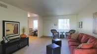 This single-floor residence offers a practical and comfortable layout designed for everyday living. The living area is spacious and carpeted, providing a soft, warm underfoot feel. Large windows allow generous amounts of natural light to fill the room, creating a bright and airy atmosphere. The neutral wall tones contribute to an open, clean aesthetic that can be easily personalized. The living space accommodates a variety of seating arrangements, as evidenced by the combination of a plush sofa, wicker accent chairs, and ottoman-style coffee tables, suggesting a versatile and inviting environment. Adjacent to the living area, the dining space transitions seamlessly via an open passageway, featuring hard flooring and room for a dining table and chairs. This open-concept connection between the two areas enhances the sense of flow and spaciousness throughout the home. A closet door is visible along one wall of the living area, offering convenient built-in storage. The textured ceiling adds a subtle architectural detail consistent throughout the space. Overall, this residence presents a functional and well-lit single-floor layout suited for comfortable, day-to-day living.