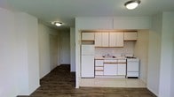 This single-floor residence offers a practical and functional layout designed for comfortable everyday living. The kitchen is a central feature of the space, equipped with ample white upper and lower cabinetry accented with warm wood trim, providing generous storage. Essential appliances include a full-size refrigerator, a gas range, and a built-in sink, all finished in white for a clean, cohesive look. The kitchen features light-toned flooring that transitions seamlessly into rich, dark hardwood-style flooring throughout the hallway and adjoining living areas. The open sightline from the kitchen into the hallway creates a sense of connected flow throughout the home. The hallway leads to additional rooms and features a closet with louvered doors, offering convenient storage access. Flush-mounted ceiling light fixtures provide consistent, practical illumination throughout. The neutral wall tones and clean finishes give the space a bright, move-in-ready feel that serves as a versatile backdrop for a variety of interior styles. Overall, the residence presents a straightforward, well-organized floor plan suited for single-occupancy or small household living.