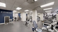 This expansive fitness center is designed to accommodate a comprehensive range of workout routines across several distinct zones. The space is finished with durable speckled rubber flooring throughout, providing a clean, professional aesthetic while ensuring safety and comfort underfoot. A drop-panel ceiling with recessed fluorescent lighting delivers consistent, even illumination across the entire floor.

The cardio zone is equipped with multiple treadmills, elliptical trainers, upright and recumbent stationary bikes, and stair-climbers, many accompanied by individual monitors. Flat-screen televisions mounted on navy blue accent walls enhance the experience. An adjacent strength-training area houses a variety of cable and selectorized machines, organized efficiently along mirrored and mural-adorned walls featuring large-scale black-and-white athletic photography.

A dedicated free weights section offers a full rack of dumbbells, adjustable benches, and a green turf-floored functional training area with additional equipment for stretching and bodyweight exercises. Structural columns are integrated cleanly into the layout without impeding movement. The entrance corridor features tasteful decorative touches including potted plants, framed artwork, and built-in shelving, setting a welcoming tone before entering the main fitness floor.