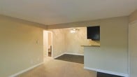 This single-floor space presents an open-concept layout that connects the living area to the kitchen through a convenient pass-through counter topped with a light-colored surface. The living area is finished with plush carpeting in a warm neutral tone, creating a comfortable underfoot feel. A ceiling fan with an integrated light fixture is centrally positioned, providing both air circulation and ambient lighting to the main living space.

The flooring transitions from carpet in the living area to a darker surface in the adjacent zone, subtly defining separate functional areas within the open plan. The kitchen is partially visible via the pass-through, where dark-finished upper cabinetry adds contrast against the warm beige walls. The walls throughout are painted in a soft, neutral beige that contributes to a warm and inviting atmosphere.

A doorway visible in the background leads to an additional room, likely a bathroom or hallway, suggesting a practical and connected floor plan. White baseboards run along the walls, providing a clean finish. Overall, the space offers a simple, functional design with cohesive neutral tones suited for everyday living.