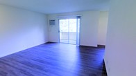 This single-floor space offers a clean, open layout highlighted by rich, dark hardwood flooring that extends throughout the main living area. The neutral, light-toned walls create a bright and airy atmosphere, complementing the natural light that streams in through a large sliding glass door. The sliding door serves as both a functional and aesthetic centerpiece, providing direct access to a private outdoor balcony and offering views of the surrounding greenery. A wall-mounted air conditioning unit is positioned near the sliding door, ensuring climate control convenience. The open layout of the living area provides flexible space for a variety of furniture arrangements. The combination of dark flooring against the light walls creates a visually appealing contrast, lending the space a modern yet understated character. The overall ambiance is clean and minimalist, making it a versatile environment well-suited for a range of uses.