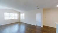 This single-floor space offers a clean, open layout with a welcoming and functional design. The main living area is defined by rich, dark hardwood flooring that contrasts beautifully with the crisp, neutral white walls and light-toned ceiling, creating a bright and airy ambiance throughout. Multiple windows along one wall allow generous amounts of natural light to fill the room, enhancing the sense of openness and space. The ceiling features a subtle textured finish, adding a modest architectural detail to the otherwise streamlined interior. White baseboards provide a clean border throughout, lending a polished, well-maintained appearance. An open doorway leads to an adjoining room, which appears to feature lighter flooring, suggesting a versatile secondary space that could serve a variety of purposes. The overall layout is thoughtfully straightforward, offering flexibility for various furnishing arrangements. The minimalist palette of whites and warm wood tones gives the space a timeless, neutral character that can easily adapt to a range of interior styles. A ceiling vent indicates a central climate control system, contributing to everyday comfort. Overall, this space presents a practical yet appealing environment with strong foundational finishes.