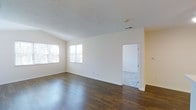 This single-floor space offers a clean, open layout with a welcoming and functional design. The main living area is defined by rich, dark hardwood flooring that contrasts beautifully with the crisp, neutral white walls and light-toned ceiling, creating a bright and airy ambiance throughout. Multiple windows along one wall allow generous amounts of natural light to fill the room, enhancing the sense of openness and space. The ceiling features a subtle textured finish, adding a modest architectural detail to the otherwise streamlined interior. White baseboards provide a clean border throughout, lending a polished, well-maintained appearance. An open doorway leads to an adjoining room, which appears to feature lighter flooring, suggesting a versatile secondary space that could serve a variety of purposes. The overall layout is thoughtfully straightforward, offering flexibility for various furnishing arrangements. The minimalist palette of whites and warm wood tones gives the space a timeless, neutral character that can easily adapt to a range of interior styles. A ceiling vent indicates a central climate control system, contributing to everyday comfort. Overall, this space presents a practical yet appealing environment with strong foundational finishes.