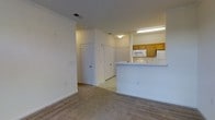 This single-floor residence showcases a practical, open-concept layout that connects the living area to the kitchen, creating a cohesive and functional space. The living area is finished with wall-to-wall carpeting in a neutral tone, providing a comfortable and warm foundation throughout. Crown molding accents the upper walls, adding a refined architectural detail to the otherwise clean, understated interior.

The kitchen is positioned toward the rear of the open floor plan and features a raised breakfast bar that serves as a natural divider between the cooking and living spaces. Warm oak cabinetry lines the upper walls of the kitchen, offering ample storage, while white appliances — including a built-in microwave, oven range, and refrigerator — provide a classic, cohesive look. Overhead fluorescent lighting ensures the kitchen area is well-illuminated.

A hallway visible from the main living space leads to additional areas of the home, flanked by multiple paneled doors that suggest closet or utility storage. The flooring transitions from tile in the kitchen and hallway to carpet in the living area. The overall color palette is light and neutral, creating a bright, welcoming ambiance throughout the space.