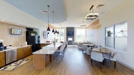 This single-story space showcases a thoughtfully designed open-concept layout that seamlessly integrates a kitchen bar area, casual dining, and a comfortable lounge. The kitchen bar features a generous marble-topped island with warm wood cabinetry, illuminated by a striking cluster of matte black cone pendant lights that draw the eye upward toward the elevated tray ceiling detail. Rich hardwood-style flooring flows continuously throughout, grounding the space with warmth.

The lounge area anchors the far end of the room, centered around a sleek, wall-mounted fireplace framed in dark stone, flanked by large windows that invite abundant natural light and offer views of the surrounding greenery. A sophisticated geometric chandelier overhead adds a modern, layered lighting element to this gathering area.

Along the right wall, an intimate dining zone accommodates multiple seating arrangements with woven accent chairs and wood-topped tables — ideal for small groups. The walls feature a subtle warm wood-tone accent band, tying together the contemporary design palette of whites, naturals, and black fixtures. Overall, the space exudes a modern yet welcoming ambiance suited for socializing and relaxation.