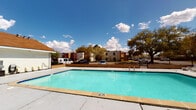 Located in Lafayette, Louisiana, this outdoor recreational facility centers around a generously sized swimming pool with clear turquoise water, surrounded by a wide concrete deck ideal for lounging. The pool area is enclosed by a sturdy black iron fence with a gated entry, ensuring controlled and secure access for residents or guests. Signage is posted at the gate, reinforcing safety guidelines for pool use.

Adjacent to the pool is a single-story pool house clad in white horizontal siding. The structure features a bold red entry door equipped with a keypad intercom system for secure access, as well as a set of elegant white French doors that provide an additional entrance point. A display window near the entry showcases festive Mardi Gras-themed artwork, reflecting the vibrant local culture of the Lafayette community.

A covered breezeway connects the pool house to the surrounding property, outfitted with a vending machine for added convenience. Decorative potted plants and barrel planters accent the entryways, adding a welcoming touch to the exterior. Mature oak trees and lush greenery are visible in the background, framing the space under the wide Louisiana sky and enhancing the overall outdoor ambiance of this well-appointed community amenity area.