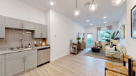 This single-floor residence features a thoughtfully designed open-concept layout that seamlessly connects the kitchen, dining, and living areas. The kitchen is outfitted with gray shaker-style cabinetry, granite countertops, a textured tile backsplash, and stainless steel appliances including a refrigerator, dishwasher, and range. Light wood-look flooring runs throughout the main living areas, complementing the bright white walls and recessed lighting overhead. Stylish globe pendant lights add a contemporary touch above the kitchen area.

The living room benefits from soaring ceilings and a glass door that invites abundant natural light while providing outdoor views through large windows dressed with sheer curtains. Two well-proportioned bedrooms feature plush carpeting and ample wall space. The full bathroom includes a tub-shower combination with tile surround, maintaining the clean, neutral aesthetic found throughout the unit. A dedicated laundry closet houses a stacked washer and dryer, adding everyday convenience. Overall, the space presents a modern, airy ambiance with cohesive finishes throughout.
