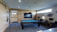This single-floor recreational space is designed with a contemporary aesthetic, combining functionality and style to create an inviting entertainment lounge. The room's most prominent feature is a sleek, dark-framed billiard table with a vibrant teal felt surface, positioned at the center and serving as the focal point of the space.

A striking wood-paneled accent wall provides a warm, natural backdrop and anchors a large wall-mounted flat-screen television, flanked by modern wall sconces that add ambient lighting. A bold, oversized black dome pendant light hangs above the billiard table, reinforcing the room's modern design sensibility.

Recessed ceiling lighting supplements the natural light that filters in through narrow horizontal windows positioned high along the walls, lending the space an open, airy quality. The flooring features wide-plank wood-look material in a cool gray tone, complementing the neutral palette of the walls and ceiling.

A contemporary sectional sofa with navy accent pillows is tucked along one wall, offering a comfortable seating area for spectators or casual lounging. A glass-paneled sliding door adds a refined, transparent architectural detail, connecting this space to an adjacent area while maintaining visual openness.