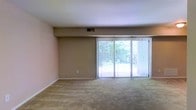 This single-floor residence offers a straightforward and functional layout designed for comfortable everyday living. The main living area is spacious and open, featuring neutral-toned carpet flooring and warm beige walls that create a calm, understated ambiance throughout the space. A prominent set of sliding glass doors spans a generous portion of one wall, fitted with vertical blinds that allow for adjustable light control. These doors serve as the focal point of the room, flooding the interior with natural light and offering a direct view of the verdant, tree-lined outdoor surroundings. The seamless connection between the interior and the greenery outside lends the space a refreshing, nature-adjacent feel. Overhead ventilation is provided via a ceiling-mounted HVAC vent, ensuring year-round climate comfort. The clean lines, neutral palette, and abundant natural light make this a versatile space well-suited to a variety of interior arrangements and personal styles. The single-floor layout adds to the ease of navigation and accessibility throughout the home.