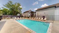 Located at 1933 Argo Rd in Conroe, Texas, this outdoor amenity area centers around a generously sized rectangular swimming pool with clear, blue-tinted water. The pool deck is finished in smooth concrete and offers ample space for relaxation, lined on multiple sides with white lounge chairs and occasional umbrella seating for shade. Entry to the pool is facilitated by built-in steps and a metal handrail on one end, providing easy access for all users. A well-maintained single-story facility building with board-and-batten gray siding and a pitched shingle roof sits adjacent to the pool, housing what appear to be restroom and utility access doors finished in a contrasting dark blue-gray. The pool area is enclosed by a black metal fence, providing both security and an open sightline to the surrounding mature trees and lush greenery. Residential structures with covered balconies are visible beyond the fence line, indicating this pool serves as a shared community amenity. Outdoor lighting poles are positioned throughout the deck area, ensuring usability into the evening hours. The overall setting conveys a well-appointed, functional outdoor retreat designed for community enjoyment under the open Texas sky.