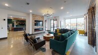 This contemporary single-floor amenity space is thoughtfully designed to accommodate a variety of social and functional activities across several distinct zones. The open-concept layout features polished concrete flooring throughout, white walls, and generous natural light streaming in through large floor-to-ceiling windows that overlook an outdoor terrace.

The main lounge area anchors the space with a striking stone-clad accent wall housing a sleek linear electric fireplace and a wall-mounted flat-screen TV. Plush teal sectional sofas and modern armchairs arranged around a live-edge coffee table create an inviting gathering spot. An elegant chandelier and pendant lighting add warmth overhead.

Adjacent to the lounge is a dining area with a large wooden communal table, patterned upholstered chairs, and a dramatic cluster of mixed-tone pendant lights hanging above. This area also benefits from direct access to the outdoor terrace through glass doors.

A dedicated media and entertainment bar area features a teal accent wall, a trio of wall-mounted flat-screen TVs set against a textured mosaic tile backsplash, and a long run of warm wood cabinetry below. A white quartz bar counter is lined with teal upholstered stools and leather-accented bar chairs.

A quieter nook near the windows offers two teal armchairs, curated artwork, and a built-in shelving unit with dark tile accents — ideal for more intimate conversation or relaxation.