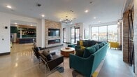 This contemporary single-floor amenity space is thoughtfully designed to accommodate a variety of social and functional activities across several distinct zones. The open-concept layout features polished concrete flooring throughout, white walls, and generous natural light streaming in through large floor-to-ceiling windows that overlook an outdoor terrace.

The main lounge area anchors the space with a striking stone-clad accent wall housing a sleek linear electric fireplace and a wall-mounted flat-screen TV. Plush teal sectional sofas and modern armchairs arranged around a live-edge coffee table create an inviting gathering spot. An elegant chandelier and pendant lighting add warmth overhead.

Adjacent to the lounge is a dining area with a large wooden communal table, patterned upholstered chairs, and a dramatic cluster of mixed-tone pendant lights hanging above. This area also benefits from direct access to the outdoor terrace through glass doors.

A dedicated media and entertainment bar area features a teal accent wall, a trio of wall-mounted flat-screen TVs set against a textured mosaic tile backsplash, and a long run of warm wood cabinetry below. A white quartz bar counter is lined with teal upholstered stools and leather-accented bar chairs.

A quieter nook near the windows offers two teal armchairs, curated artwork, and a built-in shelving unit with dark tile accents — ideal for more intimate conversation or relaxation.