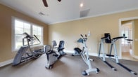 The fitness center is a single-floor space designed to support an active lifestyle, combining functional equipment with a comfortable and well-maintained environment. The room features warm beige walls accented by classic crown molding, lending a polished, residential feel to the workout space. Carpeted flooring runs throughout, providing a softer surface underfoot compared to traditional gym flooring. The ceiling is fitted with a ceiling fan, ensuring adequate air circulation during workouts, while recessed lighting supplements the natural light that enters through a window on one wall. The window also offers a pleasant outdoor view, adding an airy, open quality to the space. The fitness center is equipped with a variety of cardio machines, including a recumbent bicycle, an elliptical cross-trainer, an upright exercise machine, and what appears to be a dip and raise station, offering users diverse workout options. Ventilation is further supported by visible HVAC vents in the ceiling. Overall, the space strikes a balance between a home-like atmosphere and a functional fitness environment, making it an inviting and practical area for daily exercise.