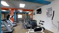 This single-floor fitness center offers a comprehensive workout environment designed to accommodate a variety of exercise needs. The space is visually energized by bold orange and teal accent beams and columns that contrast against neutral gray and white walls, creating a motivating atmosphere for users. Recessed panel lighting in the drop ceiling ensures the entire floor is evenly and brightly illuminated throughout the day, supplemented by natural light streaming in through a window along one wall. The flooring features a durable, multi-tone carpet tile suitable for high-traffic fitness use. Cardio equipment, including elliptical trainers and an exercise bike, is arranged across the main floor, while a dedicated strength training zone toward the rear houses a multi-station cable machine along with free weight benches and racks. A flat-screen television is wall-mounted at a comfortable viewing height, allowing members to stay entertained during workouts. The layout is thoughtfully organized to allow clear movement between equipment zones, providing a functional and efficient exercise experience. The overall design blends practicality with an energetic visual identity, making this fitness center a well-rounded facility for daily training.