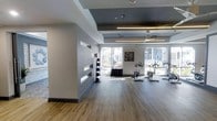 This single-floor fitness space combines modern design with functional layout, creating an inviting environment for exercise. The room features durable hardwood-style flooring throughout, providing a clean and practical surface well-suited to physical activity. A coffered ceiling with contrasting dark and light panels adds architectural interest overhead, while a large ceiling fan and pendant lighting ensure the space remains well-lit and comfortable during use.

Natural light is a defining feature of this fitness area, with expansive floor-to-ceiling windows along the exterior wall allowing generous sunlight to pour in and offering views of the surrounding outdoor greenery. The open, airy feel is further enhanced by the room's generous proportions.

A structural column divides the space subtly, with horizontal slot windows cut into the adjacent partition wall adding visual depth without compromising the open feel. A medicine ball storage rack is positioned near this partition, keeping equipment organized and accessible. Stationary cycling bikes are arranged toward the windowed wall, taking full advantage of the natural light and outdoor views. The overall ambiance is clean, contemporary, and welcoming.