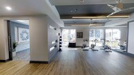 This single-floor fitness space combines modern design with functional layout, creating an inviting environment for exercise. The room features durable hardwood-style flooring throughout, providing a clean and practical surface well-suited to physical activity. A coffered ceiling with contrasting dark and light panels adds architectural interest overhead, while a large ceiling fan and pendant lighting ensure the space remains well-lit and comfortable during use.

Natural light is a defining feature of this fitness area, with expansive floor-to-ceiling windows along the exterior wall allowing generous sunlight to pour in and offering views of the surrounding outdoor greenery. The open, airy feel is further enhanced by the room's generous proportions.

A structural column divides the space subtly, with horizontal slot windows cut into the adjacent partition wall adding visual depth without compromising the open feel. A medicine ball storage rack is positioned near this partition, keeping equipment organized and accessible. Stationary cycling bikes are arranged toward the windowed wall, taking full advantage of the natural light and outdoor views. The overall ambiance is clean, contemporary, and welcoming.
