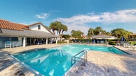 Located in the heart of Boca Raton, Florida, this community pool area at Arbor Club Way offers a resort-style outdoor retreat that embodies the relaxed elegance of South Florida living. The centerpiece is a generously sized, crystal-clear swimming pool with gentle entry steps and stainless steel railings, set atop an expansive paver-stone sundeck that provides ample space for lounging and socializing. Surrounding the pool, an array of blue lounge chairs and shaded umbrella seating areas invite residents to unwind in comfort. Towering palm trees frame the space, lending a tropical atmosphere while providing natural shade throughout the day. The adjacent clubhouse structure features a white exterior with a terracotta-tiled roofline, and a covered patio area extends the usable outdoor space for gatherings. Neatly trimmed hedges and manicured landscaping define the perimeter, adding to the polished, well-maintained character of the area. With an open-sky setting and abundant Florida sunshine, this outdoor amenity space captures the vibrant, leisure-oriented lifestyle that defines the Arbor Club community in Boca Raton.