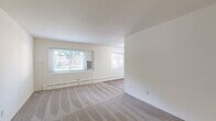 Located at 1431 Burke Ave NE in Grand Rapids, Michigan, this one-bedroom, one-bathroom unit offers a practical and efficiently organized single-floor layout. The space is divided into a living room, bedroom, kitchen, bathroom, and connecting hallway, providing a clear and functional flow throughout.

The living room is a generously sized, open area finished with neutral-toned carpet and crisp white walls. Two windows allow natural light to filter in, offering views of the surrounding neighborhood's mature trees. A window-unit air conditioner and baseboard heating system provide year-round climate control. Two entry doors — one interior and one exterior — are visible from the living area.

The galley-style kitchen features white cabinetry accented with warm wood trim, a double-basin sink with laminate countertops, a gas range, dishwasher, and refrigerator. Light wood-style vinyl flooring runs underfoot, and overhead lighting keeps the space well-illuminated. A hallway connects the kitchen to the rest of the unit.

The spacious primary bedroom includes carpet flooring, a sliding-door closet with built-in shelving, a window for natural light, and baseboard heating. The bathroom features white subway tile wainscoting, wood-toned upper cabinetry, vinyl plank flooring, and a grab bar for added accessibility.
