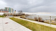 Located at 33 Ontario St in Kingston, Ontario, this outdoor Waterfront BBQ Area offers a well-appointed communal gathering space directly adjacent to the waterfront. The area features a spacious concrete patio equipped with BBQ facilities, a large dining table with seating, and additional lounge chairs positioned to take full advantage of the sweeping views of Lake Ontario. Manicured green lawns frame the patio on multiple sides, providing a pleasant natural buffer between the building and the water's edge. A classic lamp post anchors the outdoor area, adding a charming, traditional character to the space. A glass and steel railing runs along the waterfront perimeter, ensuring safety while preserving unobstructed sightlines across the lake. The surrounding landscape includes a multi-storey residential building with balconies overlooking the grounds, and nearby heritage landmarks—including what appears to be a docked historic vessel—add distinctive local character to the setting. Paved walkways connect the BBQ area to the broader waterfront promenade, making it an accessible and inviting outdoor amenity. The overall ambiance is relaxed and scenic, ideal for outdoor dining and social gatherings with panoramic waterfront views as a backdrop.