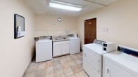 This shared amenity space is divided into two functional areas: a laundry room and a storage locker corridor, both designed to serve the practical needs of residents. The laundry room is outfitted with multiple coin-operated washing machines and dryers, including Whirlpool units, arranged for efficient use. A built-in utility sink with cabinetry above provides a convenient handwashing or pre-treatment station. The room features light tile flooring, fluorescent overhead lighting, and neutral cream-colored walls, creating a clean and functional environment. Posted signage encourages considerate use of the shared space. Adjacent to the laundry area, a carpeted corridor houses a row of individual storage lockers, each secured with its own door and hardware. The lockers are uniform in design, finished in white, and lined along one wall of the hallway. Overhead fluorescent lighting illuminates the corridor clearly. A wall-mounted utility panel is also present near the entrance of the locker area. The entry to the facility features a solid wood-toned door with a window panel, and a small framed artwork adds a modest decorative touch to the otherwise utilitarian space. Overall, this facility is straightforward, well-organized, and purpose-built for everyday residential convenience.