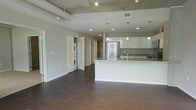 This contemporary single-floor space showcases a thoughtfully designed open-concept layout that seamlessly connects the living area to the kitchen. The kitchen is a standout feature, boasting glossy white cabinetry, a large white waterfall-edge island with an integrated sink, stainless steel refrigerator, and dark stone tile backsplash. Pendant lighting hangs elegantly above the island, complementing the recessed lighting throughout. The ceiling draws immediate attention with its exposed concrete and visible ductwork, lending an industrial character that contrasts beautifully with the clean, modern finishes below. Rich, dark hardwood flooring runs throughout the open living space, adding warmth and a sense of continuity. A dedicated in-unit laundry area is conveniently tucked behind a doorway, housing a stacked washer and dryer. Additional doorways lead to further rooms, suggesting a well-organized and functional floor plan. The neutral gray walls and abundance of recessed lighting create a bright, airy atmosphere throughout, making this a versatile and stylish space suited for modern living.