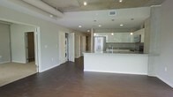 This contemporary single-floor space showcases a thoughtfully designed open-concept layout that seamlessly connects the living area to the kitchen. The kitchen is a standout feature, boasting glossy white cabinetry, a large white waterfall-edge island with an integrated sink, stainless steel refrigerator, and dark stone tile backsplash. Pendant lighting hangs elegantly above the island, complementing the recessed lighting throughout. The ceiling draws immediate attention with its exposed concrete and visible ductwork, lending an industrial character that contrasts beautifully with the clean, modern finishes below. Rich, dark hardwood flooring runs throughout the open living space, adding warmth and a sense of continuity. A dedicated in-unit laundry area is conveniently tucked behind a doorway, housing a stacked washer and dryer. Additional doorways lead to further rooms, suggesting a well-organized and functional floor plan. The neutral gray walls and abundance of recessed lighting create a bright, airy atmosphere throughout, making this a versatile and stylish space suited for modern living.