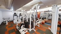 This fitness center is a fully outfitted exercise facility designed to support a wide range of workout routines. The space features a clean, modern aesthetic with a drop-panel ceiling equipped with bright recessed lighting that ensures the entire floor is well-illuminated. Large mirrors line the walls, enhancing the sense of openness and allowing users to monitor their form during workouts. The flooring consists of a durable, multi-toned carpet tile in a bold orange and dark pattern, providing both comfort and a vibrant visual energy throughout the space. The strength training area is anchored by a multi-station cable and weight machine system with substantial weight stacks, ideal for a variety of resistance exercises. Additional selectorized machines are visible throughout, catering to different muscle groups. The cardio section includes treadmills equipped with individual monitors for an engaging workout experience. Stability and exercise balls are also available, supporting flexibility and core training routines. An accent wall in a warm terracotta tone adds a welcoming pop of color to the space. Overall, the fitness center presents a well-organized, professional environment suitable for both beginner and experienced fitness enthusiasts.